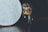 A small, stylized figurine resembling a character in wizard attire stands on a dark surface, holding a wand. The figurine has circular glasses and short hair. In the foreground, a spherical object, resembling a textured moon or a glowing orb, emits a soft light.