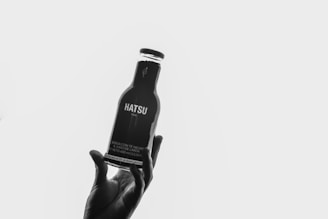 A hand holds a bottle labeled Hatsu, against a plain white background. The bottle is dark and has text on it, suggesting it contains a beverage. The composition highlights the contours of the hand and bottle in silhouette.