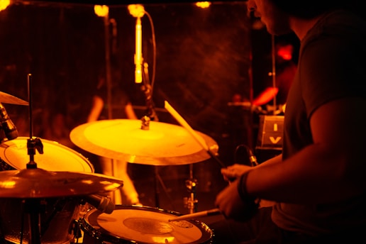 A drummer is playing a drum set on stage under warm, ambient lighting. The scene includes cymbals prominently positioned and a microphone nearby, suggesting a live music performance. There's a soft, reddish glow illuminating the musician and surroundings.