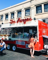 A vibrant red food truck is parked in front of an urban building, offering various items for sale as displayed on the menu boards. A woman in a polka dot dress is standing at the service window, likely placing an order. Nearby, a man is seated at a small table enjoying his meal in the outdoor setting. The truck features an eye-catching logo and signage, suggesting it specializes in South American cuisine.