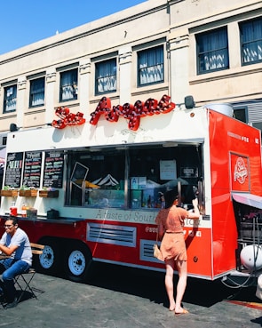 A vibrant red food truck is parked in front of an urban building, offering various items for sale as displayed on the menu boards. A woman in a polka dot dress is standing at the service window, likely placing an order. Nearby, a man is seated at a small table enjoying his meal in the outdoor setting. The truck features an eye-catching logo and signage, suggesting it specializes in South American cuisine.