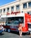 A vibrant red food truck is parked in front of an urban building, offering various items for sale as displayed on the menu boards. A woman in a polka dot dress is standing at the service window, likely placing an order. Nearby, a man is seated at a small table enjoying his meal in the outdoor setting. The truck features an eye-catching logo and signage, suggesting it specializes in South American cuisine.
