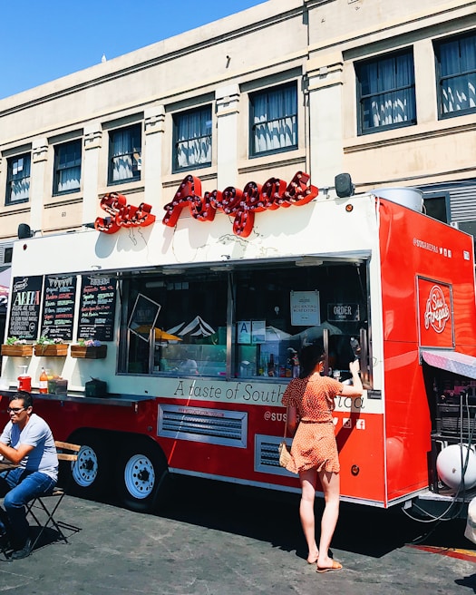 A vibrant red food truck is parked in front of an urban building, offering various items for sale as displayed on the menu boards. A woman in a polka dot dress is standing at the service window, likely placing an order. Nearby, a man is seated at a small table enjoying his meal in the outdoor setting. The truck features an eye-catching logo and signage, suggesting it specializes in South American cuisine.