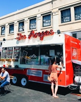 A vibrant red food truck is parked in front of an urban building, offering various items for sale as displayed on the menu boards. A woman in a polka dot dress is standing at the service window, likely placing an order. Nearby, a man is seated at a small table enjoying his meal in the outdoor setting. The truck features an eye-catching logo and signage, suggesting it specializes in South American cuisine.