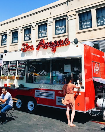 A vibrant red food truck is parked in front of an urban building, offering various items for sale as displayed on the menu boards. A woman in a polka dot dress is standing at the service window, likely placing an order. Nearby, a man is seated at a small table enjoying his meal in the outdoor setting. The truck features an eye-catching logo and signage, suggesting it specializes in South American cuisine.