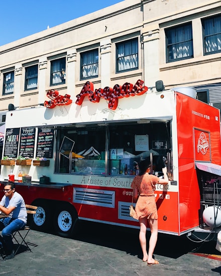 A vibrant red food truck is parked in front of an urban building, offering various items for sale as displayed on the menu boards. A woman in a polka dot dress is standing at the service window, likely placing an order. Nearby, a man is seated at a small table enjoying his meal in the outdoor setting. The truck features an eye-catching logo and signage, suggesting it specializes in South American cuisine.