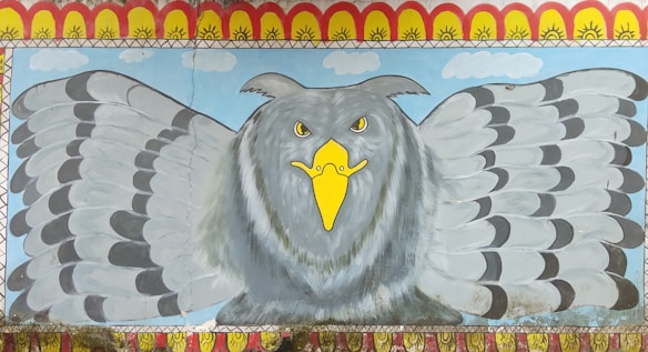 A mural featuring a large, stylized grey eagle with a prominent yellow beak and piercing eyes. The eagle's wings are spread wide, and it is set against a blue sky backdrop with small clouds. The border of the mural has a decorative pattern with yellow and red elements resembling suns.