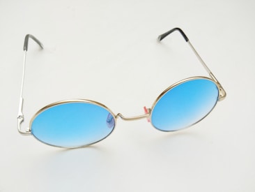 A pair of round sunglasses with thin, metallic frames and gradient blue lenses. The lens color transitions from a deep blue at the top to a lighter shade at the bottom. The frame is minimalistic with a sleek design, and the arms of the glasses are thin and metallic with black tips.