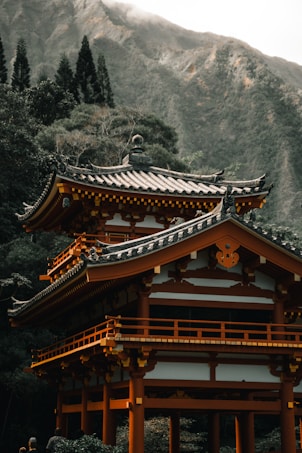 A traditional Japanese-style pagoda, characterized by its intricate architecture and sloped roofs, is set amidst lush greenery with a backdrop of towering mountains. The design features ornate details and a warm, earthy color palette.