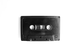 A black audio cassette tape with visible reels and tape, set against a plain white background. The cassette features small screws at the corners and a textured surface.