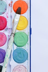 An artist's paint palette with vibrant watercolor paints in a variety of colors, including orange, yellow, green, blue, purple, red, and pink. A used paintbrush with a wooden handle rests diagonally across the palette, showing signs of colorful paint residues.