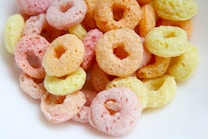 A pile of colorful, ring-shaped cereal pieces in pastel shades of pink, orange, and yellow is arranged closely together. The texture appears crunchy and porous, typical of breakfast cereals that are often enjoyed with milk.