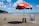 A sandy beach with a red beach umbrella labeled 'Itaipava' shading a striped blue and white beach chair and a black stool. There are a few pigeons on the sand nearby. In the background, the ocean extends to the horizon with several cargo ships visible. A green hillside is to the left under a partly cloudy blue sky.