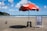 A sandy beach with a red beach umbrella labeled 'Itaipava' shading a striped blue and white beach chair and a black stool. There are a few pigeons on the sand nearby. In the background, the ocean extends to the horizon with several cargo ships visible. A green hillside is to the left under a partly cloudy blue sky.