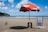 A sandy beach with a red beach umbrella labeled 'Itaipava' shading a striped blue and white beach chair and a black stool. There are a few pigeons on the sand nearby. In the background, the ocean extends to the horizon with several cargo ships visible. A green hillside is to the left under a partly cloudy blue sky.