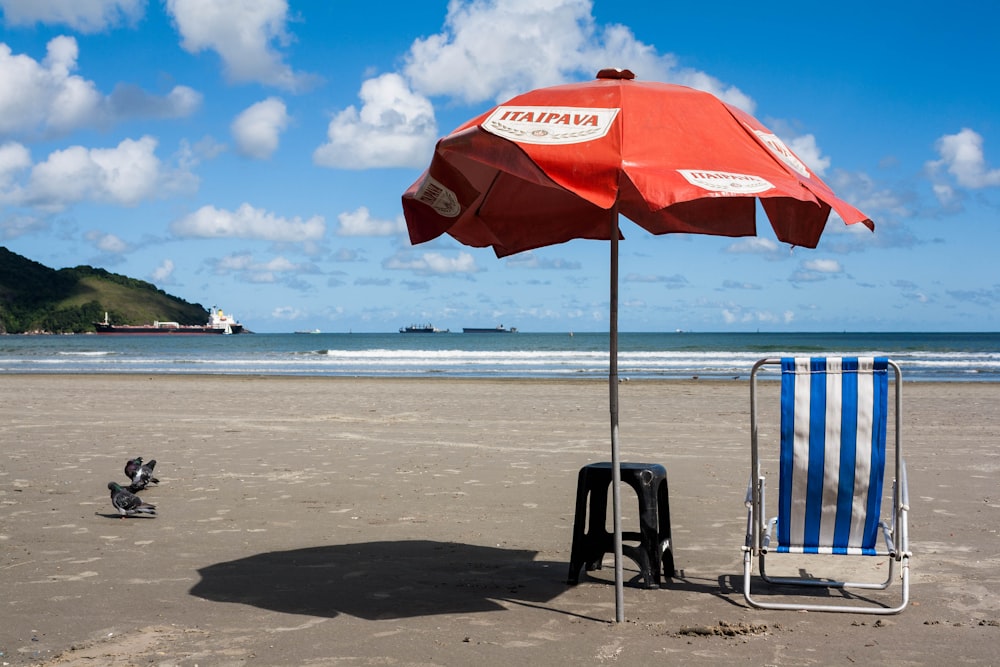 Roter und weißer Sonnenschirm in der Nähe von blauem und weißem Liegestuhl und schwarzem Plastikhocker am Strand unter weißem und blauem Himmel