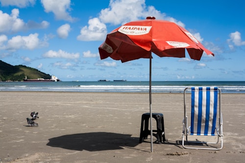 A sandy beach with a red beach umbrella labeled 'Itaipava' shading a striped blue and white beach chair and a black stool. There are a few pigeons on the sand nearby. In the background, the ocean extends to the horizon with several cargo ships visible. A green hillside is to the left under a partly cloudy blue sky.