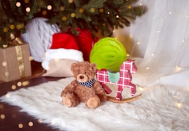 A cozy holiday scene featuring a plush teddy bear wearing a blue scarf with white stars, sitting next to a red and black plaid rocking horse ornament on a fluffy white rug. Behind them, a Christmas tree with lights is partially visible, along with a gift wrapped in brown paper and gold ribbon, a Santa hat, and a green decorative paper honeycomb ball.