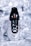 A single sneaker with a sleek, modern design lies on a metallic, crumpled background. The shoe features a white base with black laces and accents, as well as orange stitching detail. The brand's name is visible on the tongue of the shoe.