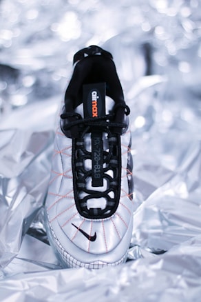 A single sneaker with a sleek, modern design lies on a metallic, crumpled background. The shoe features a white base with black laces and accents, as well as orange stitching detail. The brand's name is visible on the tongue of the shoe.