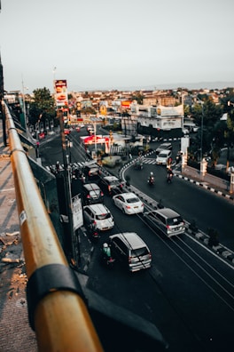 An aerial view of a busy urban intersection with several cars and motorcycles navigating through multiple lanes. Tall buildings line the street, and traffic lights are visible. Billboards and advertisements, including a prominent one featuring a burger, contribute to the bustling city atmosphere. The sky is clear, providing ample natural light.