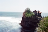 A coastal scene featuring a traditional Balinese temple perched on a rocky cliff overlooking the ocean. Greenery surrounds the temple, and waves crash against the rocks below. The temple has characteristic ornate architecture with pointed roofs and intricate carvings.