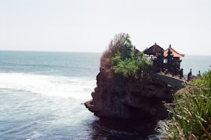 A coastal scene featuring a traditional Balinese temple perched on a rocky cliff overlooking the ocean. Greenery surrounds the temple, and waves crash against the rocks below. The temple has characteristic ornate architecture with pointed roofs and intricate carvings.