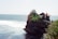 A coastal scene featuring a traditional Balinese temple perched on a rocky cliff overlooking the ocean. Greenery surrounds the temple, and waves crash against the rocks below. The temple has characteristic ornate architecture with pointed roofs and intricate carvings.