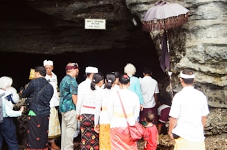 A group of people dressed in traditional clothing gather around the entrance of a cave. A sign above reads 'HOLY SPRING WATER AIR SUCI'. An umbrella with fringe is positioned beside the cave opening. The crowd appears to be engaged in a ritual or ceremony.