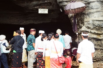 A group of people dressed in traditional clothing gather around the entrance of a cave. A sign above reads 'HOLY SPRING WATER AIR SUCI'. An umbrella with fringe is positioned beside the cave opening. The crowd appears to be engaged in a ritual or ceremony.