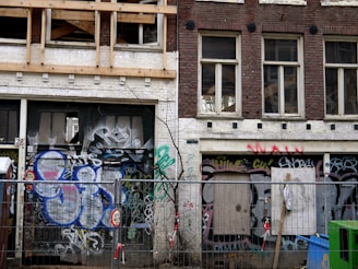 An urban scene featuring two old brick buildings with boarded-up windows and doors. The facades are heavily covered in graffiti, with a mix of colors and styles. A construction barrier with warning signs is in the foreground, indicating that the area might be under renovation or restricted access.