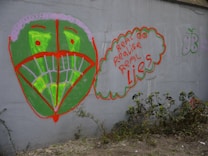 A graffiti art piece on a gray wall featuring a large, stylized green face with a red outline. The eyes and mouth are highlighted in bright green and red. To the right, a thought bubble containing the phrase 'real eyes realize real lies' in red and orange lettering. Nearby, there is a small butterfly shape outlined in green. In front of the wall, some plants are growing in the soil.