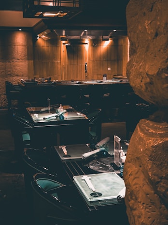 Dimly lit restaurant interior with rows of neatly arranged tables set with glass placemats, rolled napkins, and glass cups. The warm lighting casts a cozy glow on the wooden walls and stone features, creating an intimate dining atmosphere.