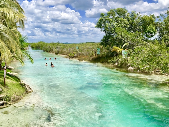 A serene natural setting with crystal-clear turquoise water flowing through a lush, green landscape. Several people are enjoying the water, surrounded by dense vegetation and trees under a bright, partly cloudy sky.