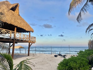 A rustic wooden hut with a thatched roof is situated on a sandy beach. Palm trees frame the view, and the calm ocean stretches to the horizon under a mostly clear blue sky with a few scattered clouds.