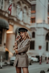 A woman poses confidently on a city street, wearing a stylish plaid blazer and a black cap. She holds a magazine with red lips on the cover, partially concealing her face. The background features an urban setting with blurred architectural elements, contributing to a fashionable and artistic atmosphere.