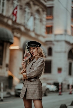 A woman poses confidently on a city street, wearing a stylish plaid blazer and a black cap. She holds a magazine with red lips on the cover, partially concealing her face. The background features an urban setting with blurred architectural elements, contributing to a fashionable and artistic atmosphere.