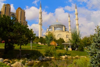 A grand mosque with multiple minarets stands prominently, surrounded by lush greenery and a tranquil pond. The sky is partly cloudy, creating a serene and peaceful atmosphere. Nearby, there is a large, modern building with intricate architecture.