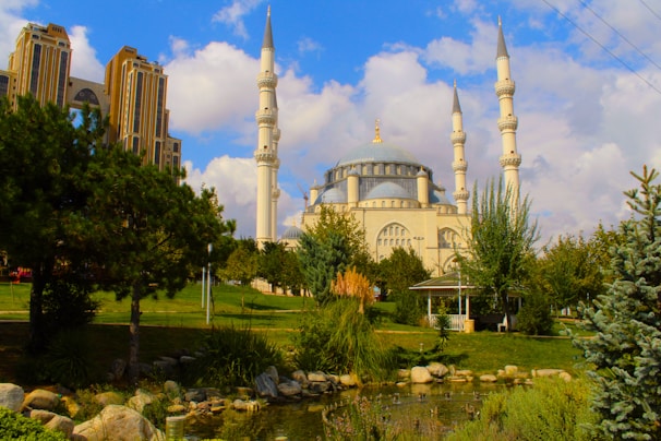 A grand mosque with multiple minarets stands prominently, surrounded by lush greenery and a tranquil pond. The sky is partly cloudy, creating a serene and peaceful atmosphere. Nearby, there is a large, modern building with intricate architecture.