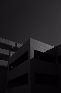 A building with a minimalist design is seen in silhouette under a stark sky. The structure has sharp angles and a geometric form, with shadows creating contrast on its surface.