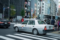 A taxi is waiting at a crossing in a busy city with multiple pedestrians on the sidewalk. The background features several tall buildings with various signs and advertisements. There are other vehicles and traffic lights in view, creating an urban street scene.