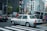 A taxi is waiting at a crossing in a busy city with multiple pedestrians on the sidewalk. The background features several tall buildings with various signs and advertisements. There are other vehicles and traffic lights in view, creating an urban street scene.