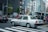 A taxi is waiting at a crossing in a busy city with multiple pedestrians on the sidewalk. The background features several tall buildings with various signs and advertisements. There are other vehicles and traffic lights in view, creating an urban street scene.
