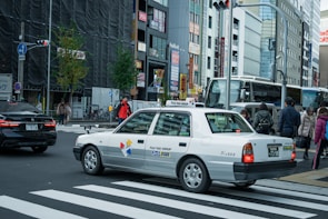 A taxi is waiting at a crossing in a busy city with multiple pedestrians on the sidewalk. The background features several tall buildings with various signs and advertisements. There are other vehicles and traffic lights in view, creating an urban street scene.