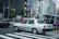 A taxi is waiting at a crossing in a busy city with multiple pedestrians on the sidewalk. The background features several tall buildings with various signs and advertisements. There are other vehicles and traffic lights in view, creating an urban street scene.