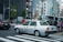 A taxi is waiting at a crossing in a busy city with multiple pedestrians on the sidewalk. The background features several tall buildings with various signs and advertisements. There are other vehicles and traffic lights in view, creating an urban street scene.