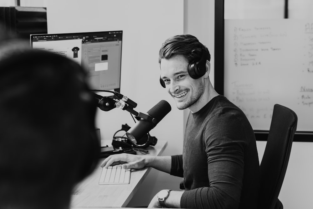 greyscale photo of man sitting by the table using headphones and microphone while smiling