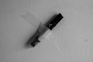 A black marker is secured to a textured surface using a piece of clear tape. The angle of the marker creates an intersection with the tape, forming an X shape. The overall tone of the image is monochrome, casting a shadow of the marker on the surface.