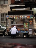 A street food cart selling various items like pretzels, hot dogs, and drinks is parked on the road. A person wearing a hooded sweatshirt is crouched in front of the cart. The cart is adorned with colorful images of the food being sold and several bottles of beverages are displayed on top.