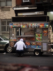 A street food cart selling various items like pretzels, hot dogs, and drinks is parked on the road. A person wearing a hooded sweatshirt is crouched in front of the cart. The cart is adorned with colorful images of the food being sold and several bottles of beverages are displayed on top.